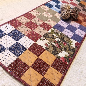May include: A patchwork table runner with a variety of floral and geometric patterns in shades of red, brown, blue, and green. The runner is quilted and has a brown border.