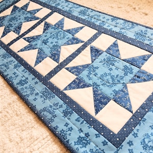 May include: A quilted table runner featuring a repeating star pattern. The stars are made of white and blue fabric, with a floral design. The runner has a light blue border with a dotted pattern and a darker blue edge.