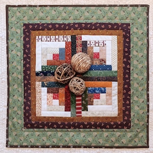 May include: A handmade quilt with a patchwork design in shades of brown, red, blue, and green. The quilt features a log cabin pattern in the center, surrounded by floral patterned borders. Three decorative woven spheres rest on the quilt.