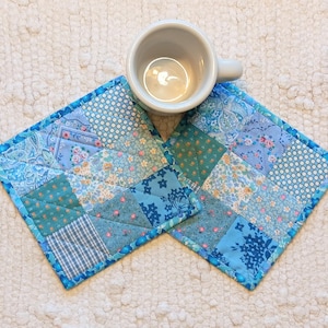 May include: Two square, patchwork coasters in shades of blue and teal, with a light blue beaded edge. A white ceramic mug sits atop one coaster. The coasters feature various floral and geometric patterns.