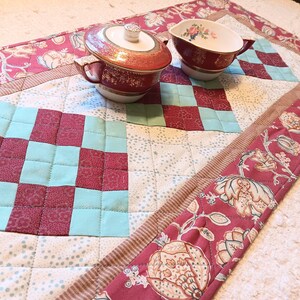 May include: A quilted table runner with a red and white floral pattern and a checkered design in shades of red and aqua. Two small, red and gold teacups with lids are on the runner.