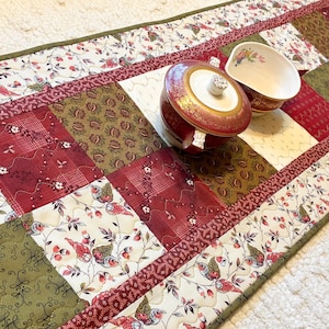 May include: A quilted table runner featuring a patchwork design of cream, olive green, and deep red fabrics with floral and scroll patterns. A round, lidded ceramic sugar bowl and a matching creamer with gold trim sit on the runner surface.