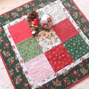 May include: A festive Christmas table runner with a patchwork design. The runner features red, green, and white squares with various patterns, including dots, snowflakes, and floral prints. A green border with cardinal birds surrounds the squares. A pinecone, ornament, and figurine are on top.