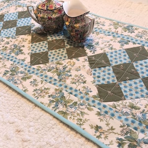 May include: A quilted table runner with a floral pattern in shades of blue, green, and cream. The runner features a patchwork design with square blocks and a light blue border. A teapot and creamer sit on top.