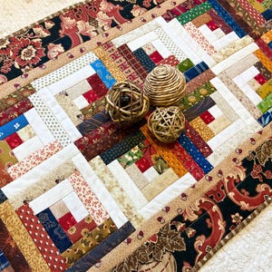 May include: A rectangular patchwork table runner featuring a log cabin quilt pattern with various multi-colored fabric strips in red, blue, green, and tan. The border displays a black fabric with a floral pattern. Three woven rattan spheres sit on top.