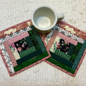 May include: Two square fabric coasters featuring a log cabin quilt pattern with strips of floral, plaid, and solid fabric in shades of pink, dark green, and cream. A plain white ceramic mug sits on top of one coaster, partially obscuring the design.