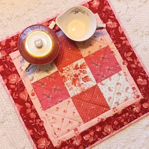May include: A red and pink quilted table runner with a floral pattern. Two porcelain teacups are on top of the runner.