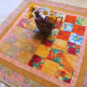 May include: A colorful patchwork quilt table runner with a floral pattern. A white pitcher with a floral design sits on top of the runner, filled with white and yellow flowers.