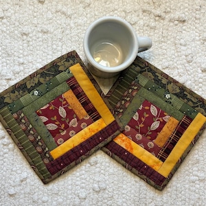 May include: Two square fabric coasters featuring a log cabin quilt pattern with strips of fabric in shades of olive green, mustard yellow, deep red, and brown with floral and plaid prints, accompanied by a plain white ceramic mug.
