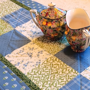 May include: A colorful teapot and teacup set with a floral and fruit design, resting on a patchwork quilt table runner. The quilt features blue, green, and white patterned squares. The teapot has a lid and a curved handle.