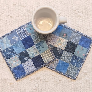 May include: Two blue and white patchwork fabric coasters with a white ceramic mug on top of one of the coasters.