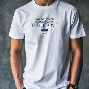 Può includere: T-shirt bianca con la scritta "need two female volunteers for a THREESOME" in un font stilizzato. Sotto il testo ci sono tre pulsanti "Subscribe" in blu, viola e lavanda. La maglietta è indossata.