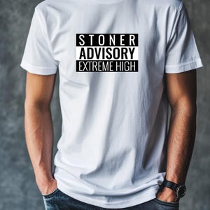 Può includere: T-shirt bianco con la scritta "STONER ADVISORY EXTREME HIGH" in un design rettangolare bianco e nero. La maglietta è bianca e ha maniche corte. La persona indossa jeans blu e un orologio nero.