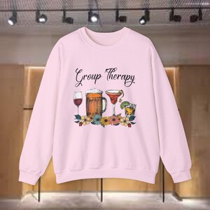 Può includere: Felpa rosa chiaro con la scritta "Group Therapy". La grafica presenta un bicchiere di vino, un boccale di birra, un cocktail e un bicchierino, circondati da fiori. Un top casual e confortevole.