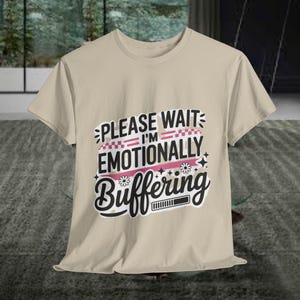 May include: A beige t-shirt with the text "PLEASE WAIT I'M EMOTIONALLY Buffering" in a playful font. The design includes pink and white accents, flowers, and a loading bar graphic. The shirt is made of a soft material.
