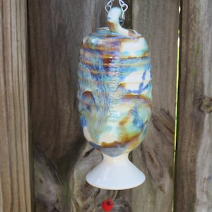 May include: A ceramic hummingbird feeder with a white base and a blue, brown, and white glazed design. The feeder is hanging from a chain.