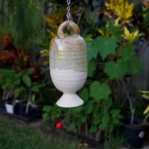 May include: A handmade ceramic hummingbird feeder with a cream-colored base and a light brown and green upper section. The feeder has a red plastic feeding tube and is suspended by a metal chain. The background features lush green foliage.