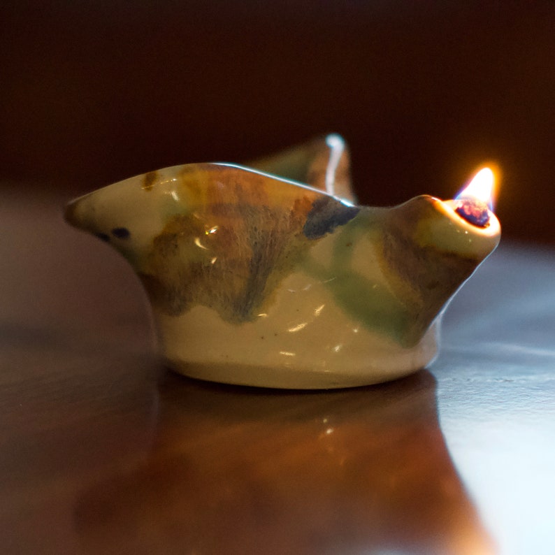 May include: A ceramic oil lamp with a brown, green, and blue glaze. The lamp is lit and the flame is visible.