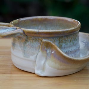 May include: Handmade ceramic shaving scuttle with a built-in bowl and handle. The scuttle features a white base with a brown and blue glaze. The bowl is designed to hold hot water for a warm, luxurious shave.