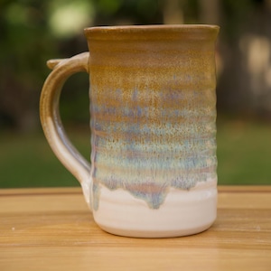 May include: A handmade ceramic mug with a tall, cylindrical shape. The mug features a gradient of colors, transitioning from a warm brown at the top to shades of blue and green in the middle, and a white base. It has a large, curved handle.