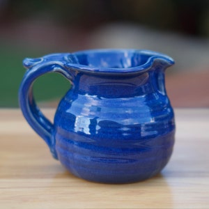 May include: A small, blue ceramic pitcher with a rounded body and a slightly flared lip. The pitcher has a handle that curves up and over the lip.