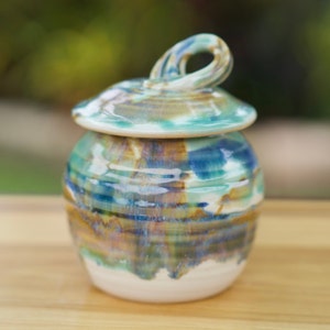 May include: A small ceramic jar with a lid and a loop handle. The jar is decorated with a blue, green, and brown glaze.