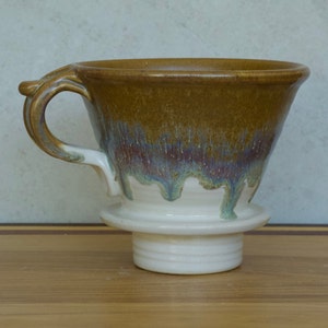 May include: A ceramic coffee pour-over with a handle. The top is brown, transitioning to blue and green, with a white base. The pour-over is designed to sit on a mug or carafe for brewing coffee.