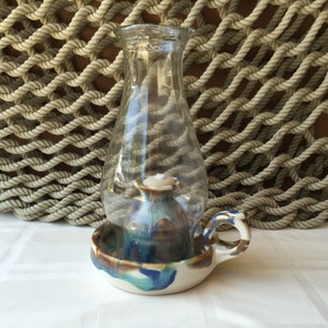 May include: A ceramic oil lamp with a blue, brown, and white glaze. The lamp has a glass chimney and a handle. The lamp is sitting on a white surface.