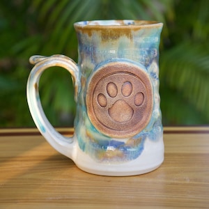 May include: A handcrafted ceramic mug with a paw print design. The mug features a textured surface with a blend of blue, green, and brown glaze. The handle is curved, and the mug has a white base. The mug is approximately 5 inches tall.