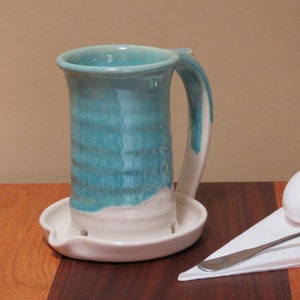 May include: A handmade ceramic mug with a turquoise glaze and a white base and handle. The mug sits on a matching white saucer with a spoon rest. The mug is approximately 6 inches tall.