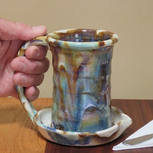 May include: A handmade ceramic mug with a unique, textured design. The mug features a handle and a built-in saucer. The colors include shades of blue, brown, and cream. The mug is held by a hand.