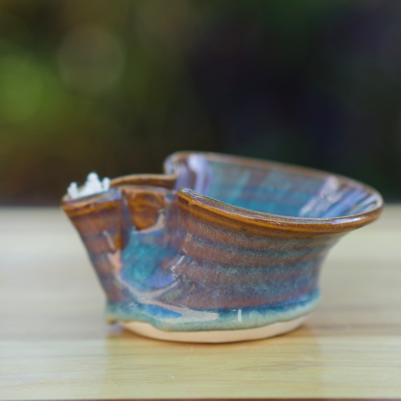 May include: A small, handmade ceramic bowl with a unique, asymmetrical shape. The bowl is glazed in shades of brown, blue, and green, creating a mottled effect. The bowl has a slightly chipped edge, adding to its rustic charm.