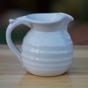 May include: A white ceramic creamer with a rounded body and a slightly flared lip. The creamer has a simple, elegant design with a smooth, glossy finish.