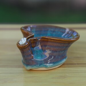May include: A small, teal and brown ceramic dish with a spout. The dish has a unique, wavy shape and a glazed finish.
