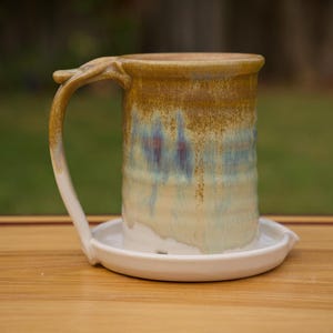 May include: Handmade ceramic mug with a unique handle design, resting on a matching saucer. The mug features a blend of earthy tones, with a gradient of brown, blue, and cream colors. The mug is approximately 5 inches tall.