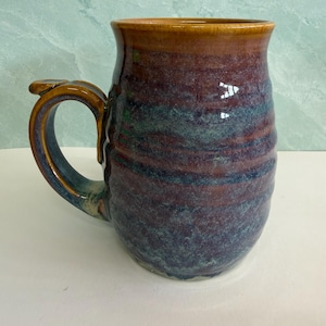 May include: A ceramic mug featuring a rounded body with a ribbed texture. The glaze displays a blend of deep burgundy, dark blue, and muted teal tones, with a solid amber-brown rim and a matching curved handle extending from the side.