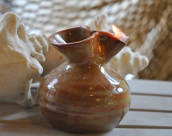 May include: A brown ceramic oil lamp with a wavy rim and a single wick burning. The lamp is sitting on a white surface.