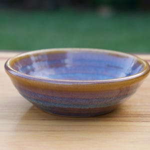 May include: A small, round ceramic bowl featuring a glossy glaze with horizontal bands of deep blue, purple, and earthy brown tones. The rim is finished in a smooth, solid mustard-yellow glaze, creating a distinct contrast against the interior.