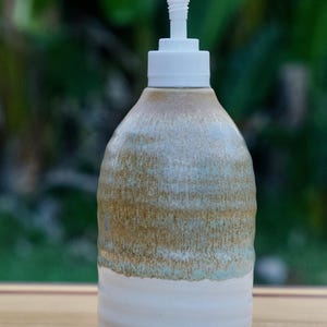 May include: A ceramic soap dispenser with a white pump. The body of the dispenser is a mix of tan, light blue, and white, with a textured, layered appearance. The bottom portion is a solid white with horizontal ridges.