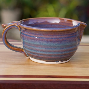 May include: A handmade ceramic mixing bowl with a handle and a pouring spout. The bowl features a blend of blue, brown, and teal hues, with a white base. The bowl is sitting on a wooden surface.