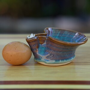 May include: A brown and blue ceramic egg cup with a brown egg inside. The egg cup has a unique shape and is glazed with a mottled finish.