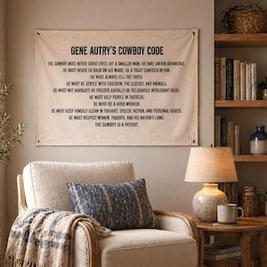 Puede incluir: Un tapiz de pared de lona con el texto "GENE AUTRY'S COWBOY CODE" en letras negras. El texto enumera principios como la honestidad, la amabilidad y el respeto. El lienzo se muestra sobre un sillón beige con un cojín estampado y una manta a cuadros.