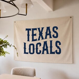 May include: A rectangular off-white canvas wall banner featuring the dark blue text "TEXAS LOCALS" in a bold, serif typeface. The banner has metal grommets in the top corners for hanging, with a clean, hemmed edge design on a plain wall surface.