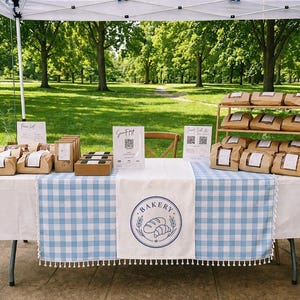Peut inclure: Un stand de boulangerie sur un marché en plein air, présentant divers produits de boulangerie dans des emballages en papier brun. La table est recouverte d'une nappe blanche et d'un chemin de table à carreaux bleus et blancs avec un logo de boulangerie.