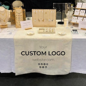 Può includere: Esposizione di gioielli, tra cui collane e orecchini, su una tovaglia bianca. Il tavolo presenta vari espositori, tra cui opzioni in legno e metallo. La tovaglia ha il testo "Your CUSTOM LOGO website.com" stampato.