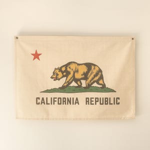 May include: A beige canvas flag with a red star in the top left corner. A brown bear walking on a green patch of grass is in the center of the flag. The text "CALIFORNIA REPUBLIC" is printed in brown at the bottom of the flag.