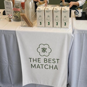 Op de afbeelding: Een wit tafelkleed met de tekst "THE BEST MATCHA" en een wolk-vormige afbeelding. Vier pakken Minor Figures Barista Oat Organic staan op de tafel. Andere items zijn een klein kattenbeeldje en een glazen bak met houten stokjes.