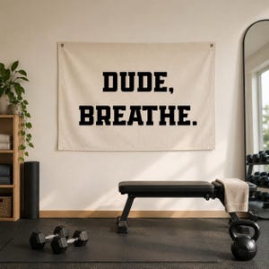 May include: A rectangular off-white fabric wall hanging featuring the black text DUDE, BREATHE. in a bold, block-style font. The banner includes metal grommets in the top corners for mounting, displaying a simple and clear typographic design.