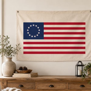 May include: A beige wall hanging featuring the American flag with 13 stars in a blue square and 13 red and white stripes. The flag is displayed on a wall above a wooden table with decorative items.