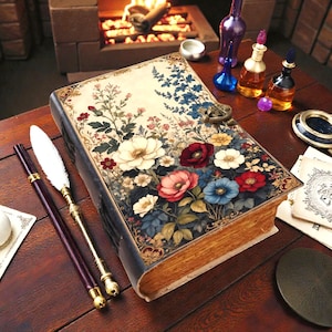May include: A vintage-style journal with a floral design featuring white, red, pink, and blue flowers. The journal has a dark blue cover and a metal clasp. Two quill pens and other decorative items are on a wooden table.